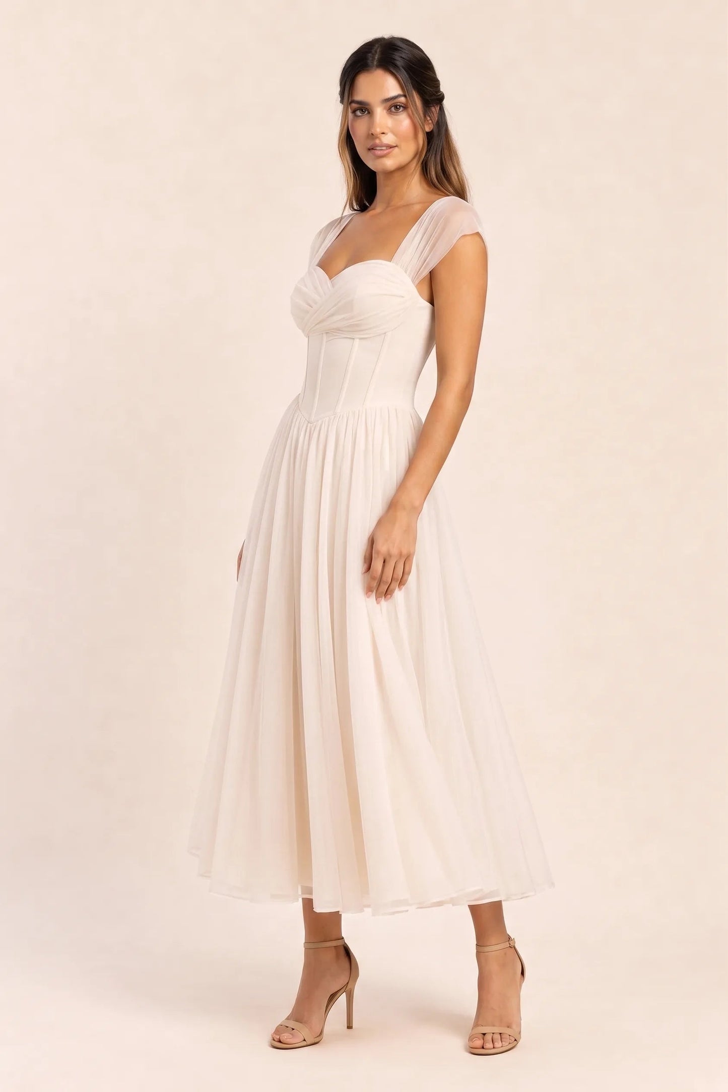 Apricot - Model wearing a romantic midi dress featuring a ruched sweetheart neckline, sheer cap sleeves, structured corset bodice with visible boning, and a full layered tulle skirt, standing indoors near a sunlit window.