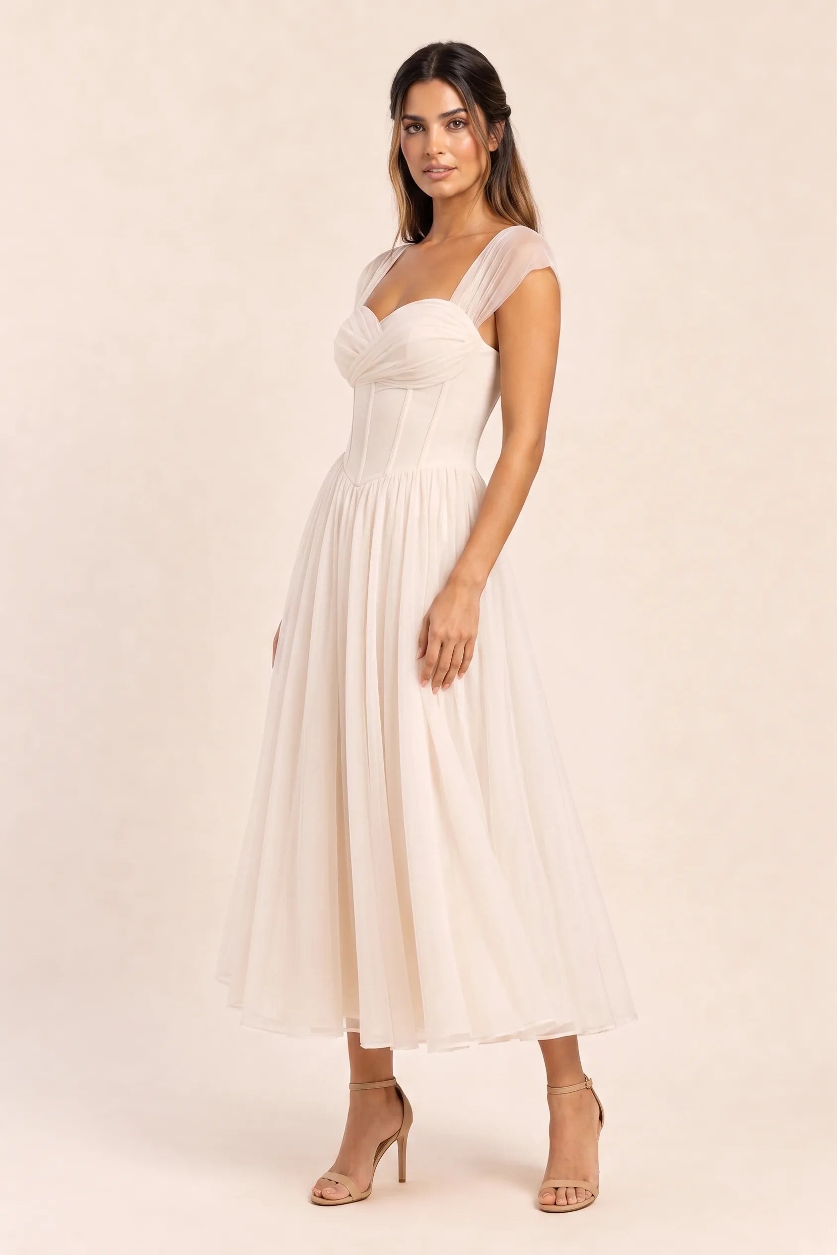 Apricot - Model wearing a romantic midi dress featuring a ruched sweetheart neckline, sheer cap sleeves, structured corset bodice with visible boning, and a full layered tulle skirt, standing indoors near a sunlit window.