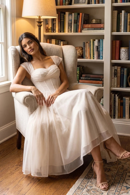 Apricot - Model wearing a romantic midi dress featuring a ruched sweetheart neckline, sheer cap sleeves, structured corset bodice with visible boning, and a full layered tulle skirt, standing indoors near a sunlit window.