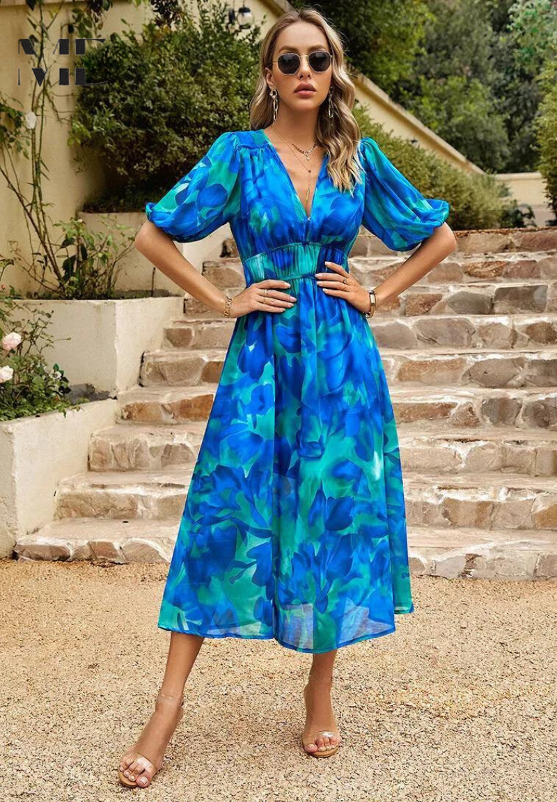 Blue - A woman wearing a vibrant, multicolored abstract floral print chiffon midi sundress with puff sleeves, a V-neckline, and a smocked waist, holding a woven straw clutch.
