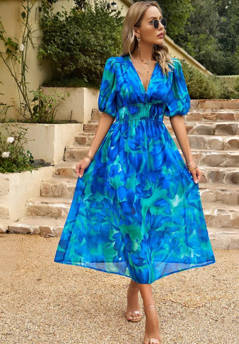 Blue - A woman wearing a vibrant, multicolored abstract floral print chiffon midi sundress with puff sleeves, a V-neckline, and a smocked waist, holding a woven straw clutch.