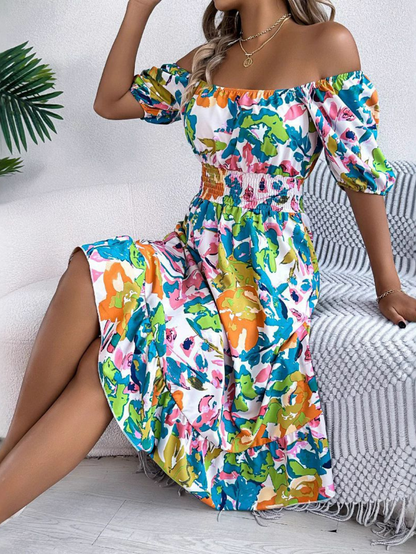 Green - A woman wearing a vibrant pink, orange, and turquoise botanical print off-the-shoulder midi sundress with a smocked waist and puff sleeves, seated on a textured white sofa.