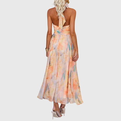 Premium - A woman wearing a sleeveless halter-neck maxi dress made of flowy watercolor chiffon in pastel orange, yellow, and blue shades, featuring a ruched waistband and a pleated A-line skirt.