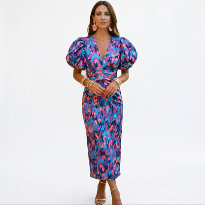 Purple - Model wearing a woven abstract pattern midi dress featuring short puff sleeves, a deep V-neckline, and a heavily ruched center bodice against a white background.