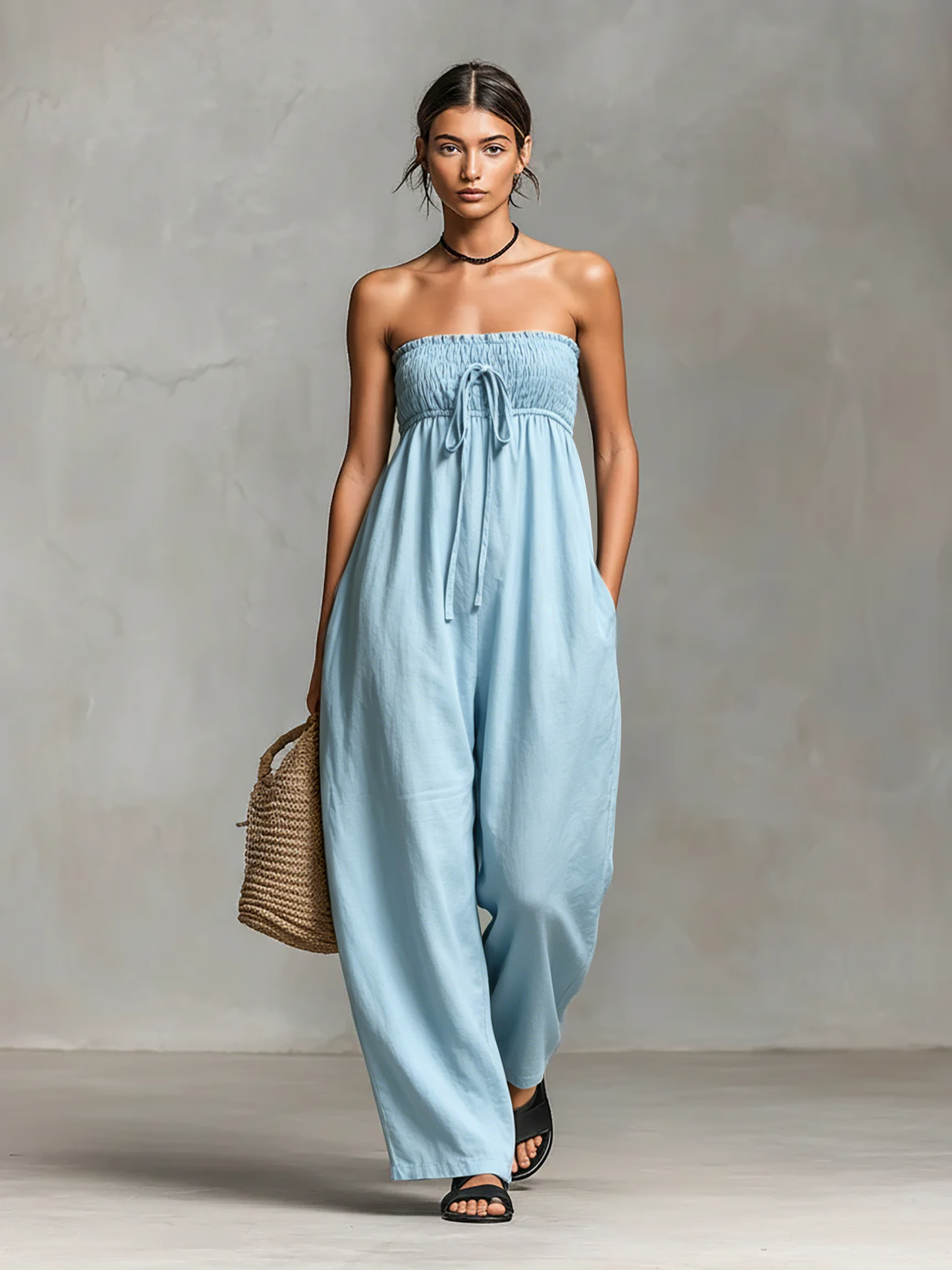 Light Blue - Model wearing a strapless wide-leg jumpsuit with a ruched bodice, front tie detail, and side pockets, holding a woven bag.